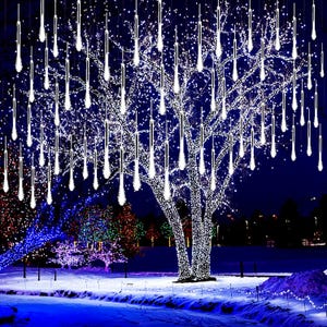 Puede incluir: Escena nocturna de un árbol decorado con luces blancas en forma de carámbanos. Las luces cuelgan hacia abajo, creando un efecto de cascada. Otros árboles en el fondo también están iluminados con luces de colores, sobre un cielo azul oscuro.
