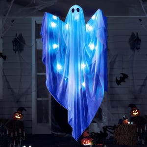 Puede incluir: Una decoración de fantasma colgante de Halloween hecha de tela azul claro con luces blancas brillantes. El fantasma tiene una cara simple con dos agujeros negros para los ojos. El fantasma está colgado frente a una casa decorada para Halloween.