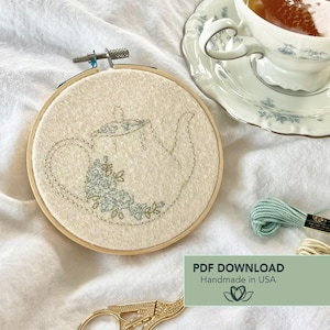May include: An embroidery hoop with a cream-colored fabric featuring a stitched teapot design with blue floral accents. A teacup and saucer are in the background, along with embroidery floss and scissors. The text "PDF DOWNLOAD Handmade in USA" is visible.