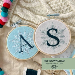 以下が含まれることがあります： 文字「A」と「S」が刺繍された2つのフープ。それぞれ雪の結晶のデザインが施されています。片方のフープは水色、もう片方はオフホワイトです。「PDF DOWNLOAD Handmade in USA」の文字が見えます。