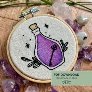 May include: Embroidered hoop art featuring a purple potion bottle with a key design. The artwork is set against a light-colored fabric, framed by a wooden hoop. The design includes black accents and star details. Text on the image reads "PDF DOWNLOAD Handmade in USA".