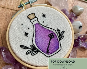 Peut inclure: Broderie sur tambour représentant une bouteille de potion violette avec un motif de clé. L'œuvre est réalisée sur un tissu clair, encadrée par un cercle en bois. Le motif comprend des accents noirs et des étoiles. Le texte sur l'image indique "PDF DOWNLOAD Handmade in USA".