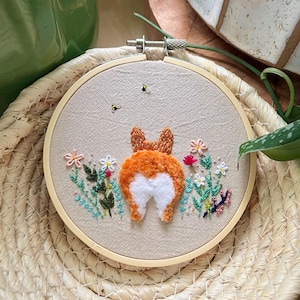 May include: Embroidered hoop art featuring a corgi's rear end, surrounded by embroidered flowers and greenery. The corgi's fluffy orange and white fur is detailed. Two small embroidered bees are above the corgi. The hoop is set in a woven basket.