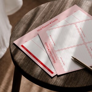 Könnte beinhalten: Zwei rosa Meal Planner und Mental Load Family Blätter mit rotem und weißem Text und einem Stift auf einem Holztisch. Die Blätter haben Abschnitte für tägliche Mahlzeiten und Aufgaben. Der Text enthält "Meal Planner Family" und "Mental Load Family".