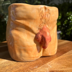 May include: A ceramic mug with a unique design, featuring a tan-colored body with a reddish-brown appendage. The mug has a textured surface and is set against a wooden background, capturing the light.