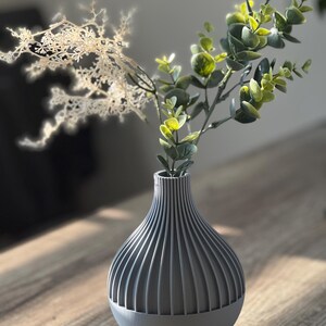 May include: A gray ceramic vase with a ribbed design, holding a sprig of green eucalyptus and a dried white flower.
