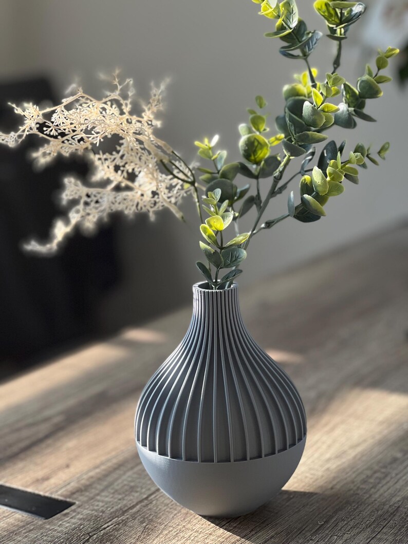 May include: A gray ceramic vase with a ribbed design, holding a sprig of green eucalyptus and a dried white flower.