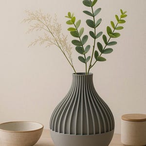 May include: A gray ceramic vase with vertical ridges, holding a bouquet of green leaves and white flowers. The vase is sitting on a wooden table.