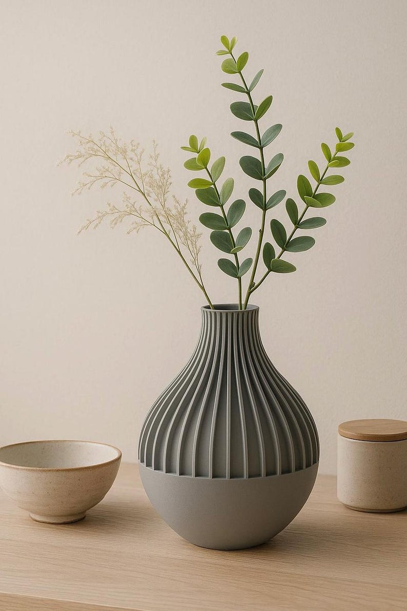 May include: A gray ceramic vase with vertical ridges, holding a bouquet of green leaves and white flowers. The vase is sitting on a wooden table.