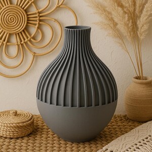 May include: A gray ceramic vase with a ribbed design sits on a woven mat. The vase is round and has a wide opening at the top. It is a modern and stylish vase that would look great in any home.