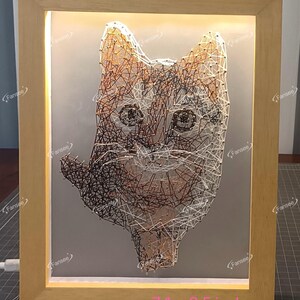 Pode incluir: Uma peça de arte de fio enquadrada que representa o rosto de um gato. O gato é feito de fio marrom, branco e laranja. A moldura é feita de madeira e tem um acabamento marrom claro. A moldura mede 7,1 polegadas por 9,5 polegadas.