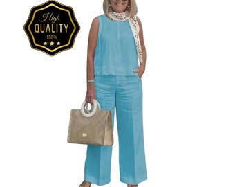May include: A sleeveless, light blue pantsuit with wide-leg pants. The outfit is paired with a beige handbag and a patterned scarf. A gold and black badge with the text "High Quality 100%" is in the upper left corner.