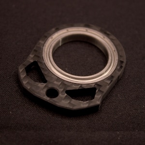 May include: A close-up shot of a carbon fibre component featuring a central circular metal bearing. The component has a textured surface and geometric cutouts. The object is set against a dark background.