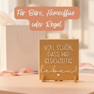 Peut inclure: Un panneau carré de couleur beige avec l'inscription blanche "VOLL SCHÖN, DASS WIR GLEICHZEITIG leben!". Le panneau est soutenu par deux petits supports beiges. Un sac cadeau blanc et un ruban rose sont en arrière-plan.