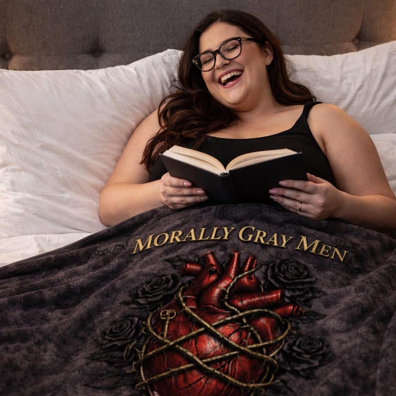 May include: A dark gray blanket with the text "MORALLY GRAY MEN" in gold lettering. The blanket features a red heart design with gold vines and black roses. The blanket is on a bed with white pillows.