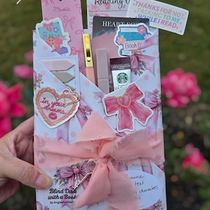 Puede incluir: Un paquete de regalo rosa y blanco con la etiqueta "Blind Date with a Book", atado con una cinta rosa y un dije de llave. El paquete contiene un bol&iacute;grafo, l&aacute;piz labial, Starbucks Via y otros art&iacute;culos relacionados con la lectura.
