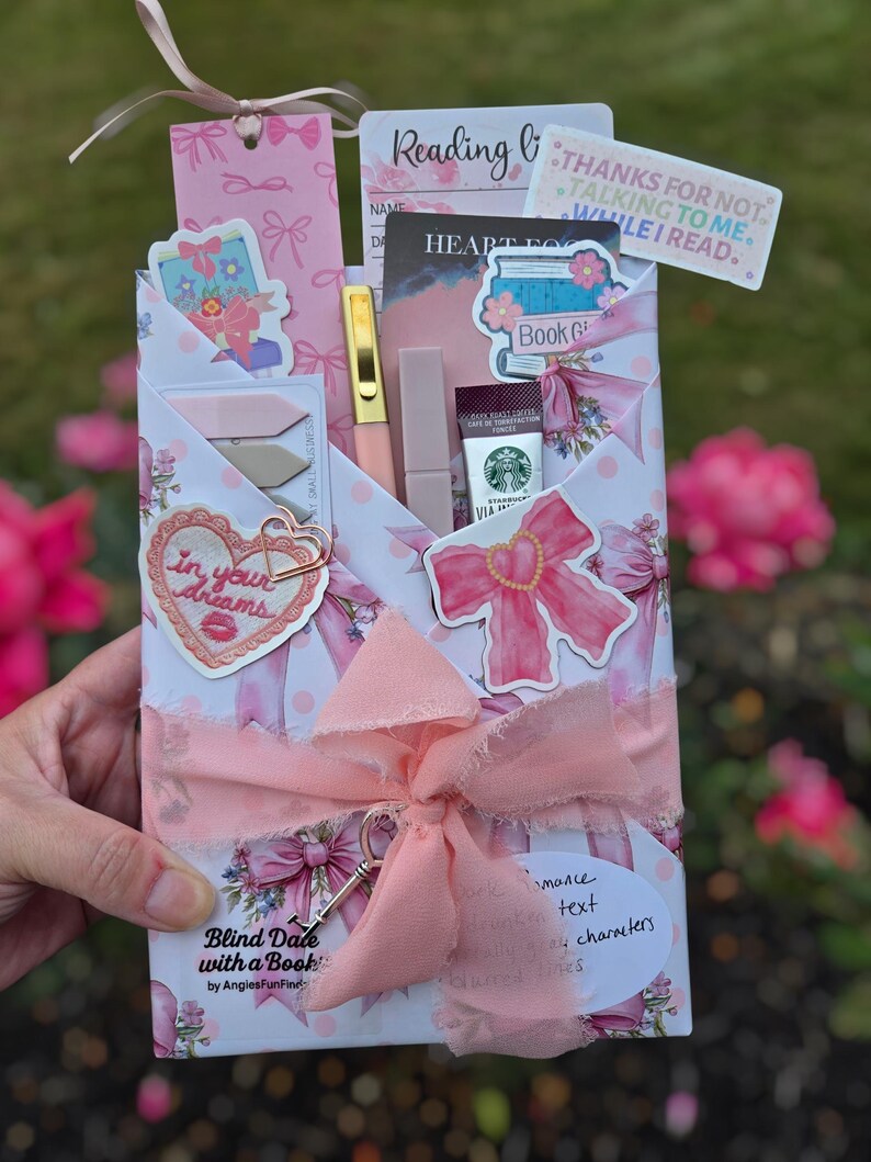 Puede incluir: Un paquete de regalo rosa y blanco con la etiqueta "Blind Date with a Book", atado con una cinta rosa y un dije de llave. El paquete contiene un bol&iacute;grafo, l&aacute;piz labial, Starbucks Via y otros art&iacute;culos relacionados con la lectura.
