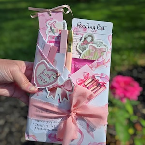 Puede incluir: Un juego de regalo rosa para un amante de los libros, envuelto en papel floral y atado con una cinta rosa. El juego incluye un marcador, pegatinas, un brillo labial y una lista de lectura. El texto en el regalo dice "book lover" y "Reading list".