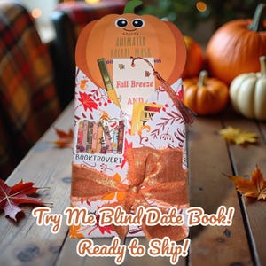 May include: A wrapped gift box with a pumpkin-shaped face mask on top. The box is decorated with fall leaves and a large orange bow. The text "Try Me Blind Date Book!" is visible on the box.