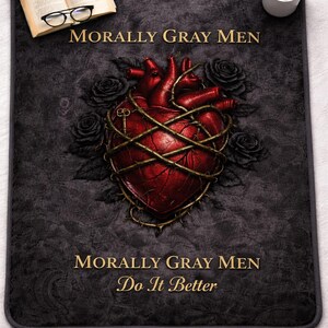 May include: A dark gray mat with a red anatomical heart design wrapped in barbed wire, surrounded by black roses. The text "MORALLY GRAY MEN" and "Do It Better" are in gold. A book, glasses, and coffee cup are also present.