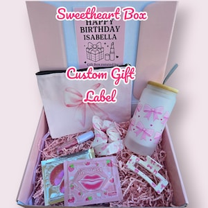 Puede incluir: Una caja de regalo rosa, etiquetada como "Sweetheart Box", llena de productos de belleza. Incluye un neceser, un vaso, una mascarilla labial, una goma para el pelo y una pinza para el pelo. La caja contiene una tarjeta que dice "Happy Birthday Isabella".