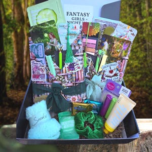 May include: A gift box filled with various items, including a face mask, socks, lip balm, and chocolates. The box contains a scrunchie, pens, and cards with the text "Fantasy Girls + Night B" and "Books of". The items are in shades of green, purple, and white.