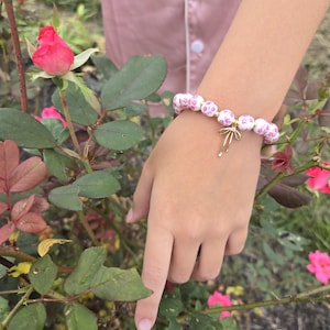 Puede incluir: Una pulsera de cuentas florales blancas y rosas con un dije dorado. La pulsera se usa en una mu&ntilde;eca cerca de un rosal con rosas rosas y hojas verdes. Las cuentas de la pulsera tienen un patr&oacute;n floral repetitivo.