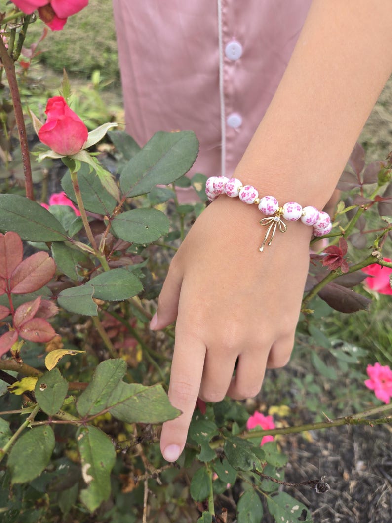 Puede incluir: Una pulsera de cuentas florales blancas y rosas con un dije dorado. La pulsera se usa en una mu&ntilde;eca cerca de un rosal con rosas rosas y hojas verdes. Las cuentas de la pulsera tienen un patr&oacute;n floral repetitivo.