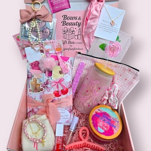 Puede incluir: Una caja de regalo rosa llena de art&iacute;culos de belleza. La caja contiene una variedad de art&iacute;culos, incluyendo un vaso con dise&ntilde;os de lazos, un collar, una pinza para el pelo y un tarro de manteca de karit&eacute; batida. La caja est&aacute; decorada con lazos y el texto "Bows & Beauty".