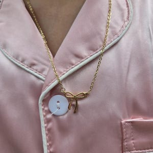 Puede incluir: Un collar dorado con un colgante de lazo. El collar se lleva sobre una camisa de pijama rosa satinada con ribete blanco y un bot&oacute;n blanco grande.