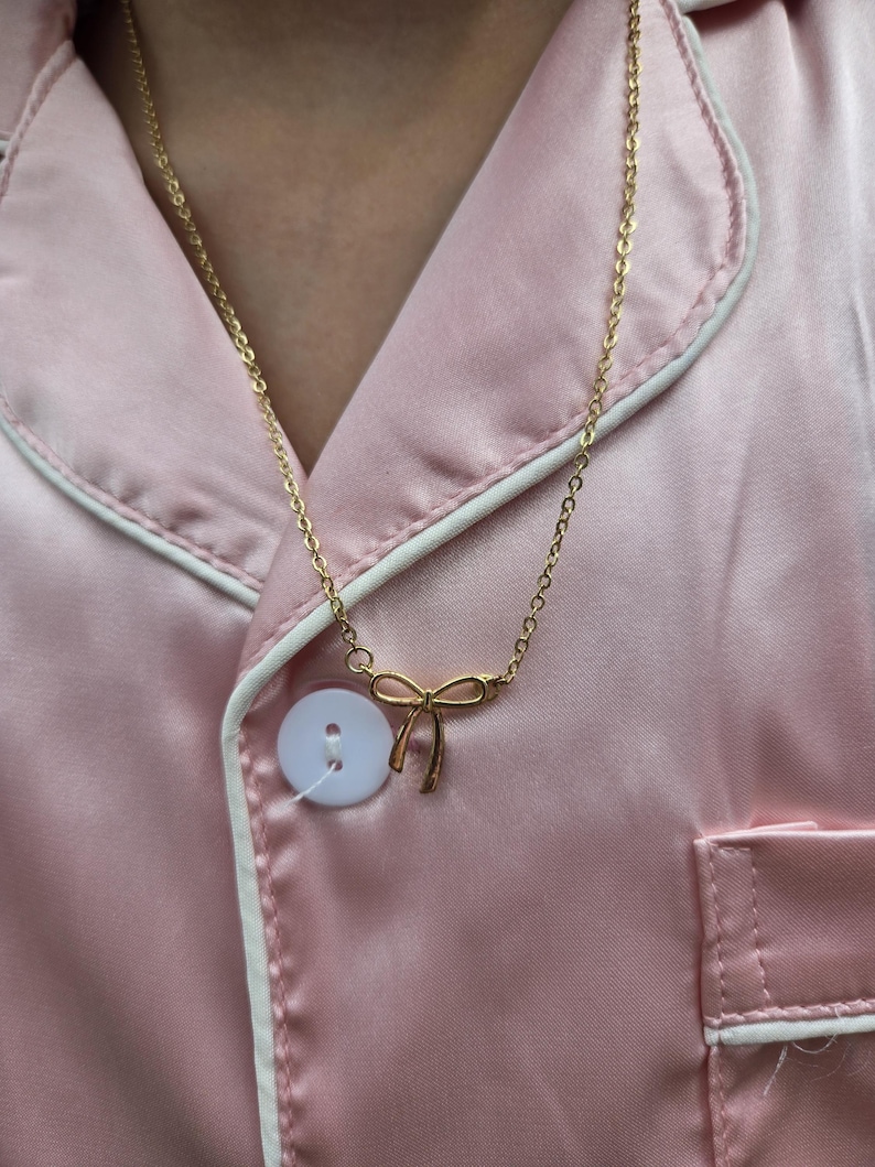 Puede incluir: Un collar dorado con un colgante de lazo. El collar se lleva sobre una camisa de pijama rosa satinada con ribete blanco y un bot&oacute;n blanco grande.