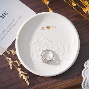 May include: A white ceramic ring dish with embossed mountain and tree designs. The dish has gold lettering with the initials "A" and "D" and a heart symbol. A silver ring with a floral design rests on the dish.