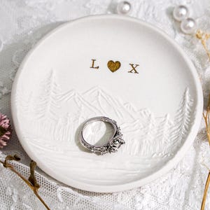 Op de afbeelding: Witte keramische ringschaal met een reliëf berg- en boomontwerp. De schaal heeft de letters "L" en "X" met een gouden hartsymbool. Een zilveren ring met een bloemenontwerp ligt in het midden van de schaal.