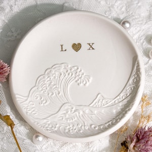 May include: Round white ceramic trinket dish with a raised wave design and gold lettering. The dish features the letters "L" and "X" with a gold heart symbol between them. The dish has a textured surface.