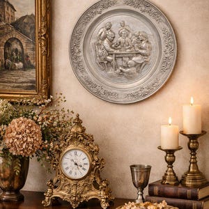 May include: A decorative wall plate with a detailed relief scene of people around a table. The image also features a gold-framed painting, a gold clock, candles, and a floral arrangement, creating a classic, elegant aesthetic.