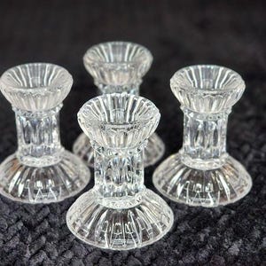 May include: Four clear glass candle holders. Each has a round base, a cylindrical stem, and a shallow, fluted top. The glass is transparent, allowing light to pass through. The candle holders are arranged on a dark, textured surface.