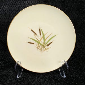May include: Cream-colored ceramic plate with a gold rim. The plate features a central design of cattails and reeds in shades of brown, green, and gold. The plate is displayed on a clear plastic stand.
