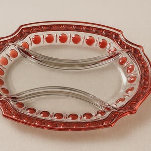 Può includere: Un piatto da portata in vetro trasparente con bordi rossi e dettagli a pois rossi. Il piatto è di forma ovale con due sezioni interne e un bordo decorativo smerlato. I dettagli rossi sono uniformemente distanziati.