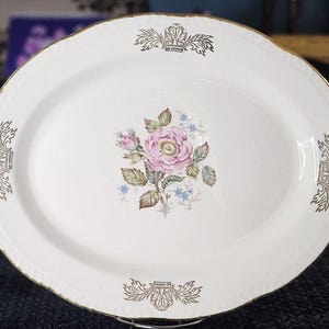 May include: Oval serving platter with a white background, featuring a central floral design of a pink rose and blue accents. The rim is trimmed with gold and decorative gold accents. A vintage piece for serving food.