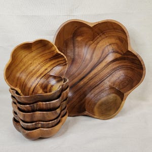 Pode incluir: Um conjunto de tigelas de madeira e uma travessa grande. As tigelas estão empilhadas e a travessa está ao fundo. O veio da madeira é visível e as tigelas têm formato de flor. A cor é marrom quente.