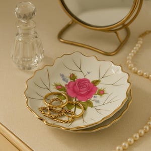 May include: A white, leaf-shaped trinket dish with a pink rose design and gold trim. Several gold rings rest inside the dish. A clear glass perfume bottle and a gold-framed mirror are in the background. A pearl necklace is also visible.