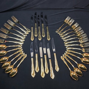 May include: A large set of gold-toned flatware arranged in two symmetrical arcs, featuring forks and spoons on the sides and a central column of knives. Each piece has a polished metallic finish with a decorative embossed pattern on the handles.