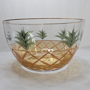 May include: A clear glass bowl featuring a gold-toned, diamond-shaped lattice pattern on the lower half and a series of green, stylized pineapple leaf motifs encircling the upper portion, finished with a thin gold rim along the top edge.