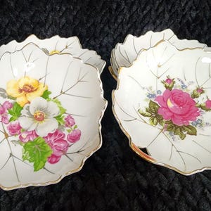 May include: Set of leaf-shaped porcelain dishes. Each dish features a white base with a gold trim and delicate floral arrangements in shades of pink, yellow, and white. The leaf design is detailed with fine gold lines.