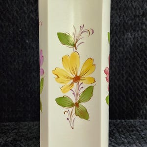 May include: Cream-colored, hexagonal vase with hand-painted floral designs. The flowers are yellow, pink, and purple with green leaves and stems. The vase has a smooth, glossy finish and is likely made of ceramic.