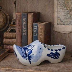 May include: A white and blue ceramic Dutch clog, with a painted floral design. The clog is displayed on a wooden shelf, with books, a globe, and a camera in the background.