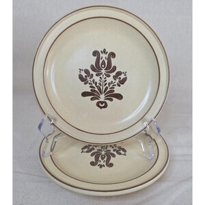 Puede incluir: Dos platos de cerámica beige con un diseño floral marrón en el centro y un borde marrón. Los platos se exhiben en un soporte de plástico transparente. El diseño floral es una flor estilizada con hojas.