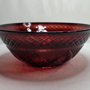 May include: A deep red glass bowl featuring a prominent diamond-cut pattern across the exterior surface. The bowl has a wide, circular opening and a tapered base, with intricate geometric detailing and starburst motifs pressed into the glass.