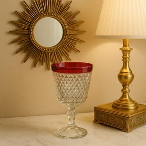 May include: A clear glass goblet with a red rim, featuring a diamond-cut pattern. The goblet is displayed on a white surface, with a gold-colored lamp and a sunburst mirror in the background. The goblet is a decorative item.