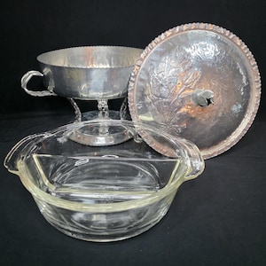 May include: A clear glass divided casserole dish with two handles, accompanied by a silver-toned metal serving stand with a hammered finish and a matching round metal lid featuring an embossed floral design and a central leaf-shaped handle.